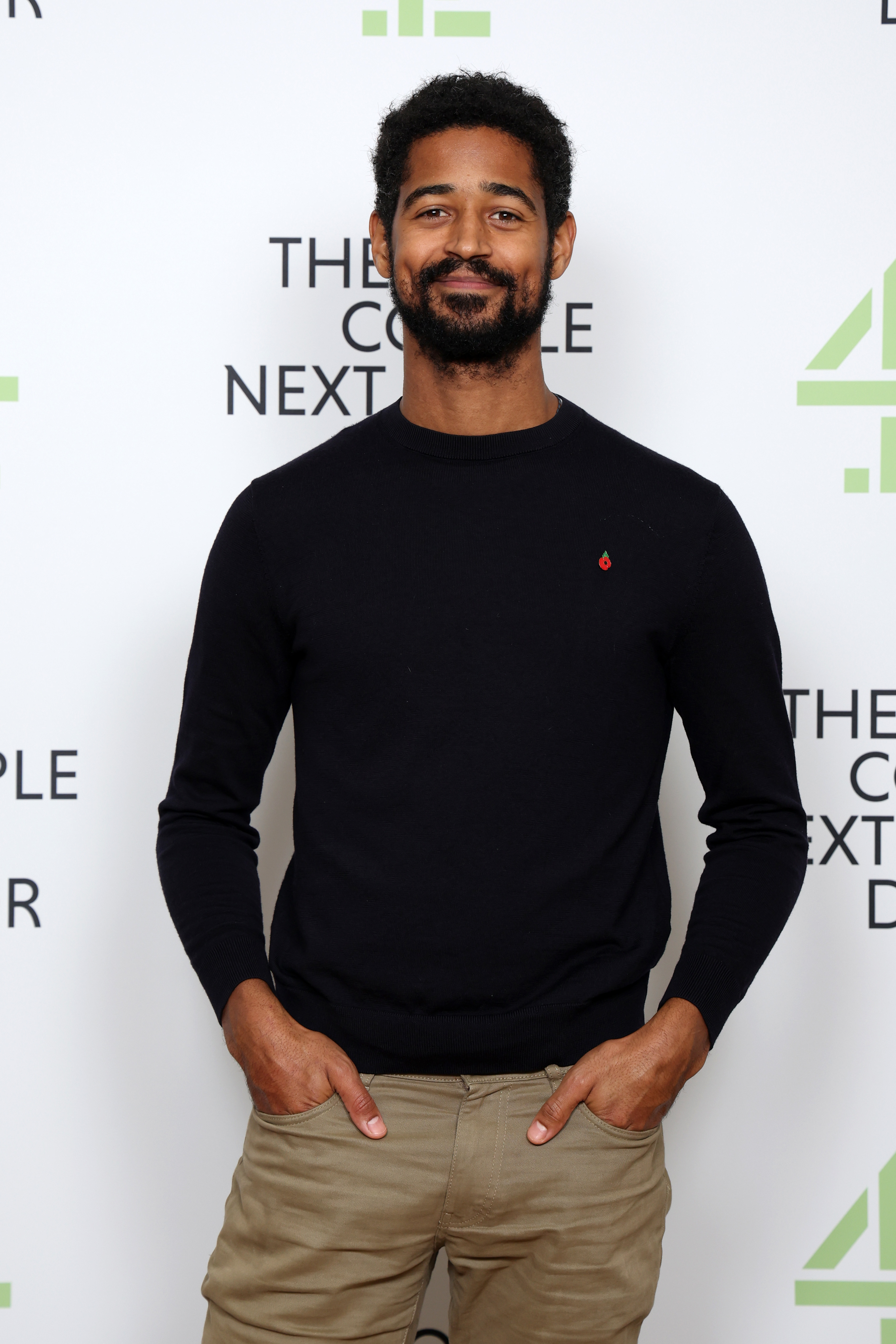 A man wearing a black sweater and beige pants standing in front of a backdrop that says "The Couple Next Door."