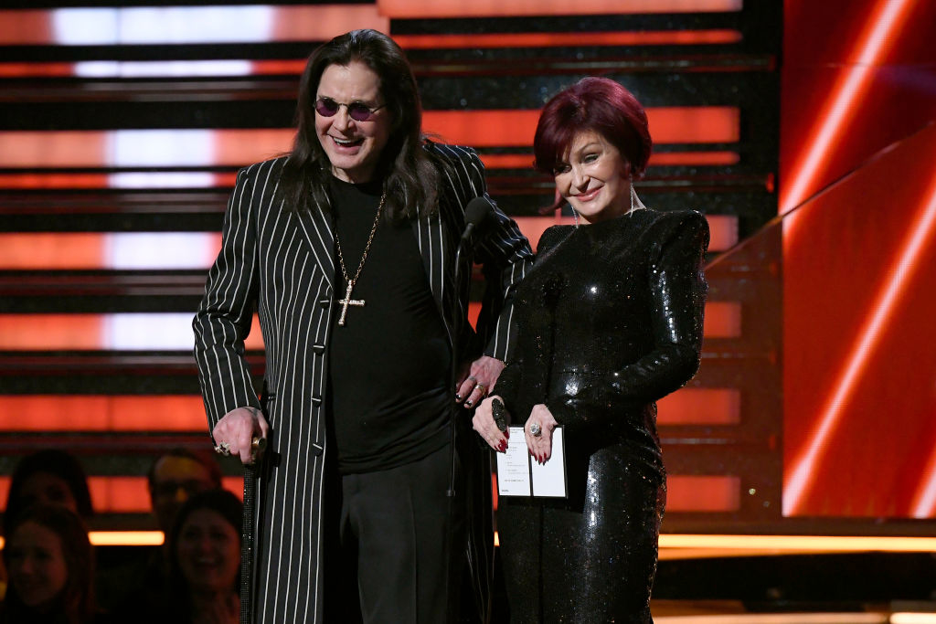 Ozzy Osbourne in a striped suit and Sharon Osbourne in a sequined dress stand on stage, smiling at an event