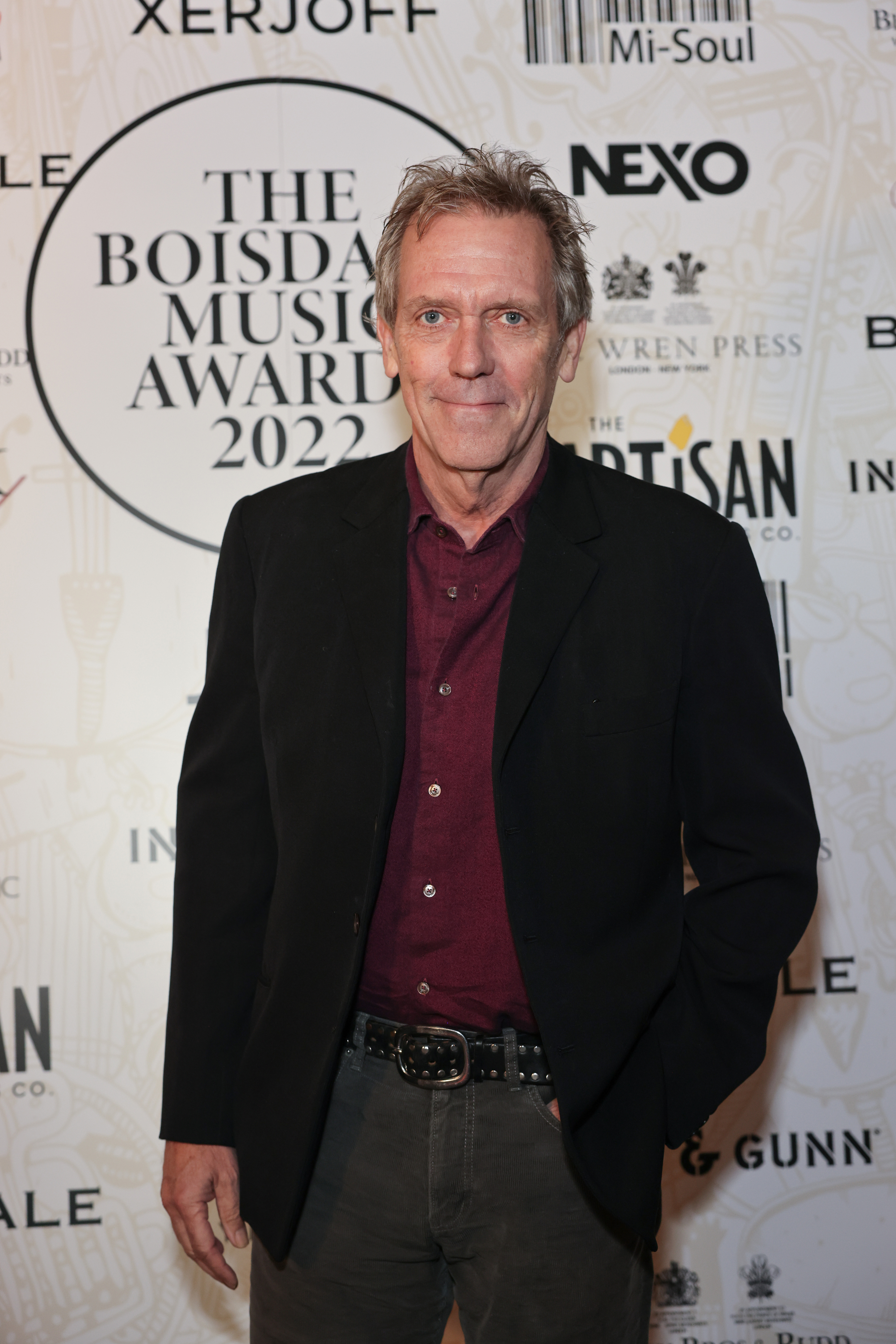 Hugh Laurie at The Boisdale Music Awards 2022, wearing a dark blazer over a red shirt, standing in front of a backdrop with event sponsors