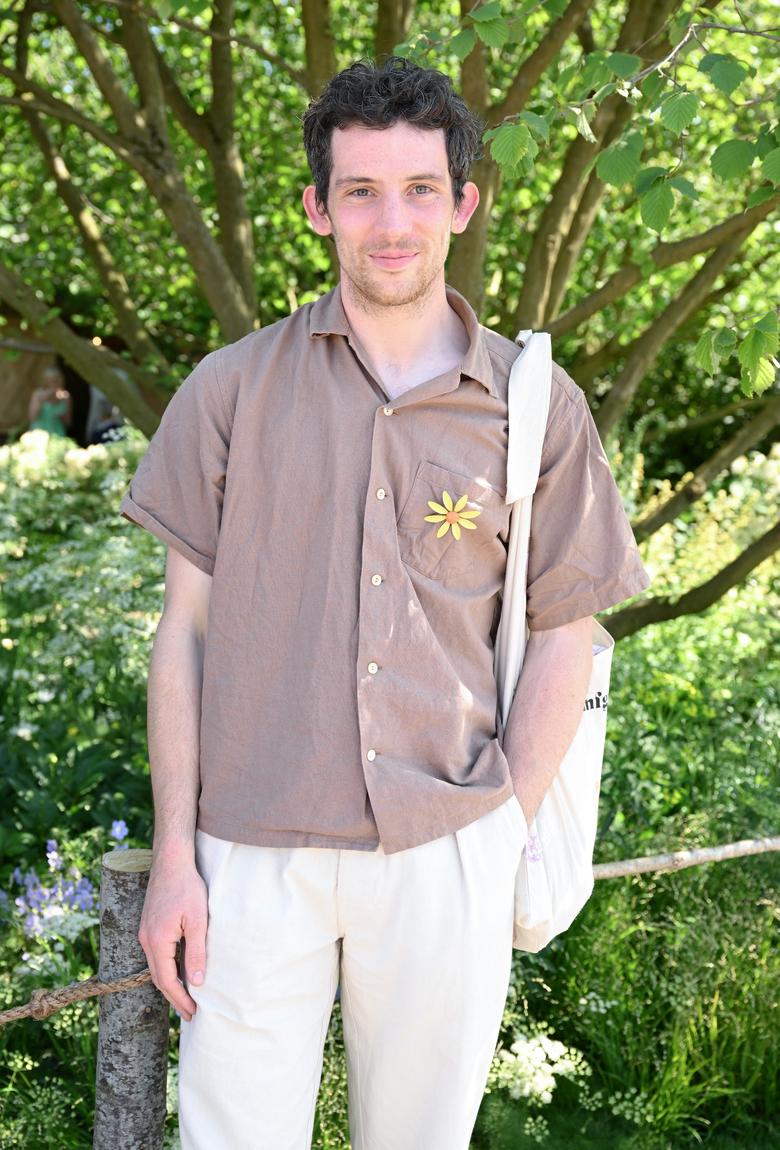 Person standing outdoors, wearing a loose button-up shirt with a flower pin and carrying a tote bag. They smile at the camera