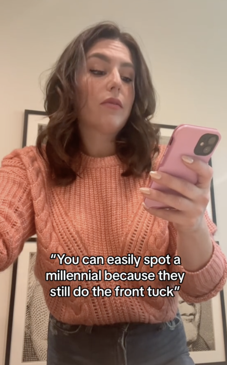 Woman in a knit sweater holds a phone and looks at it, with text below her reading, "You can easily spot a millennial because they still do the front tuck."