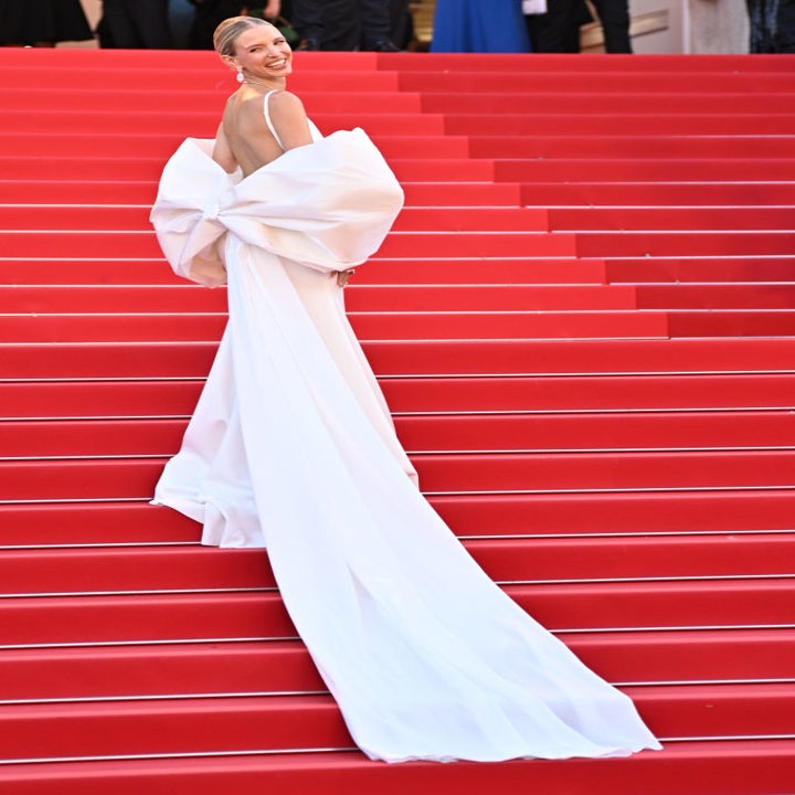 A person poses on a red carpet wearing an elegant, flowing gown with a large bow detail on the back