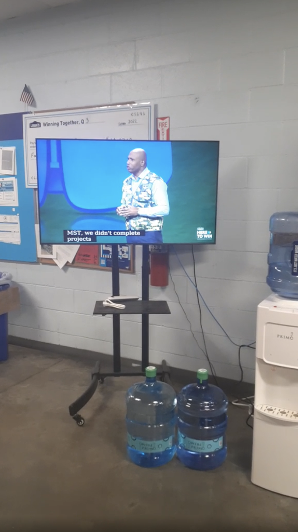 A person is speaking on a TV screen in a room with water coolers and office equipment. The screen displays subtitles but the person's name is not visible