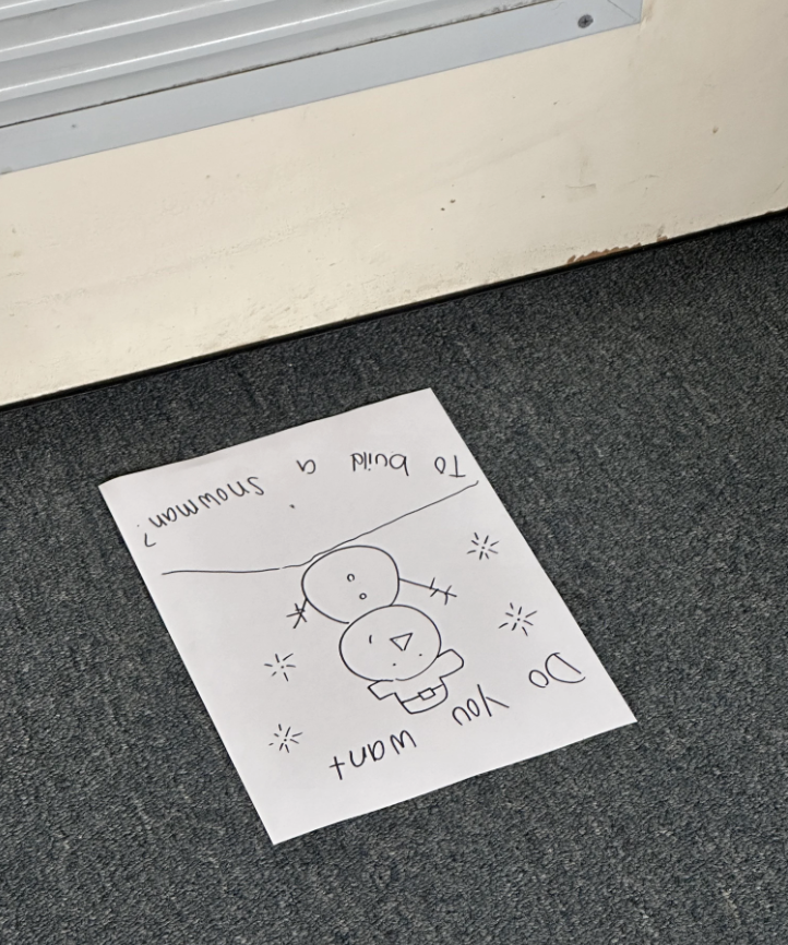 Hand-drawn image of two snowmen and the text "Do you want to build a snowman?" on a piece of paper lying upside down on a gray carpet