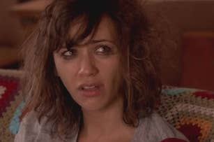Rashida Jones sitting on a couch with a concerned expression, wearing a casual shirt. The couch has a colorful crocheted blanket