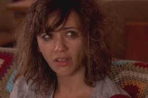 Rashida Jones sitting on a couch with a concerned expression, wearing a casual shirt. The couch has a colorful crocheted blanket