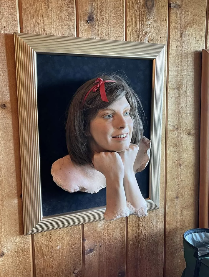 3D bust in a golden frame on a wooden wall, depicting a woman's head and hands resting under her chin with a red bow in her hair