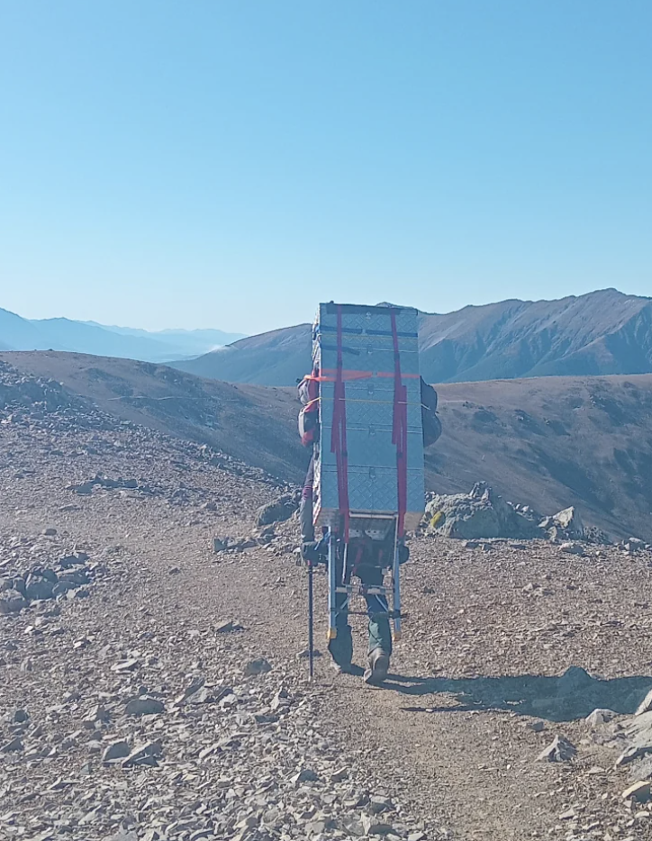 Back view of a person walking up a rocky mountain trail, carrying a large metal crate strapped to their back. Rugged mountains are visible in the background