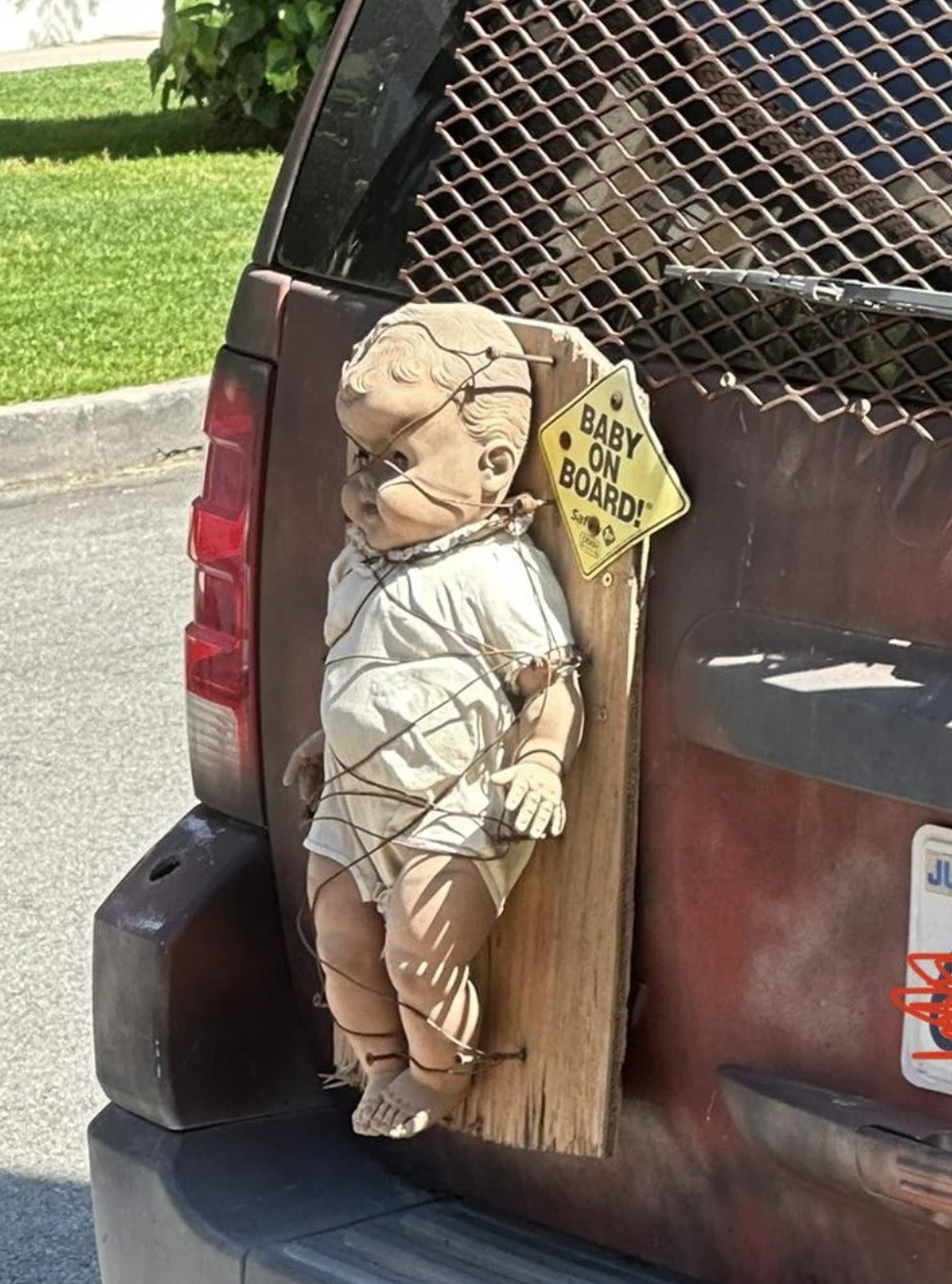A worn-out creepy baby doll is tied to a wooden plank on the back of a car. A "Baby on Board" sign is next to it