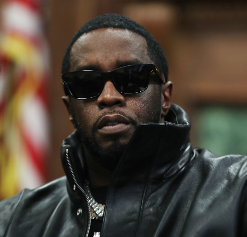 Sean "Diddy" Combs wearing sunglasses and a leather jacket, looking directly at the camera. An American flag is blurred in the background