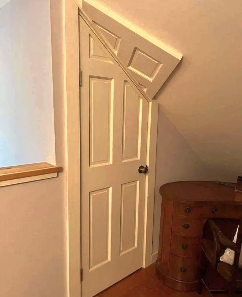 A door with a triangular cutout at the top to fit under a sloped ceiling, next to a brown wooden desk and chair