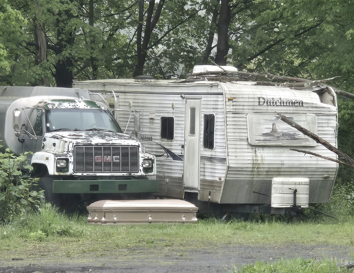 An old camper with the word "Dutchmen" is parked near a green truck; a metallic object and tree branches are on the camper's roof. A coffin lies on the ground in front