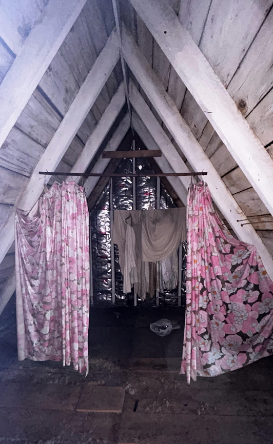 An attic space with A-frame wooden planks and floral print curtains. Laundry hangs from a line, and the room appears rustic and unfinished