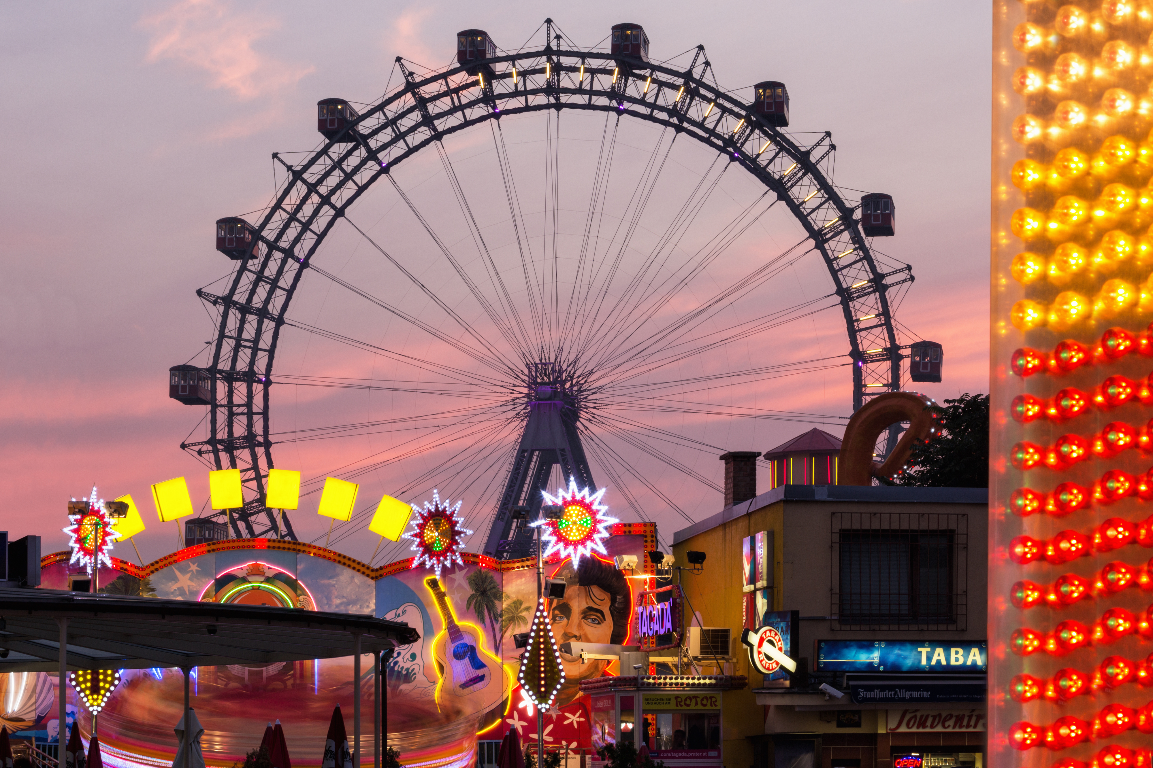 A lively carnival scene with a large ferris wheel, various illuminated attractions, and booths at sunset