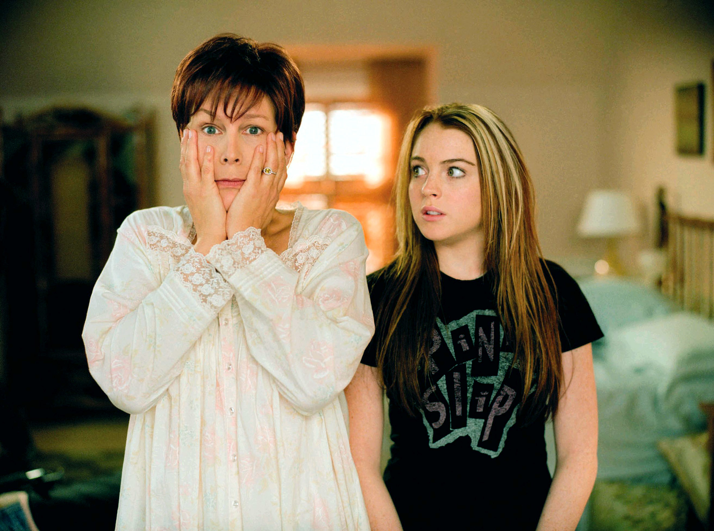 Jamie Lee Curtis with her palms on her face wearing pajamas next to Lindsay Lohan looking at confused in a band tee in a scene from "Freaky Friday"