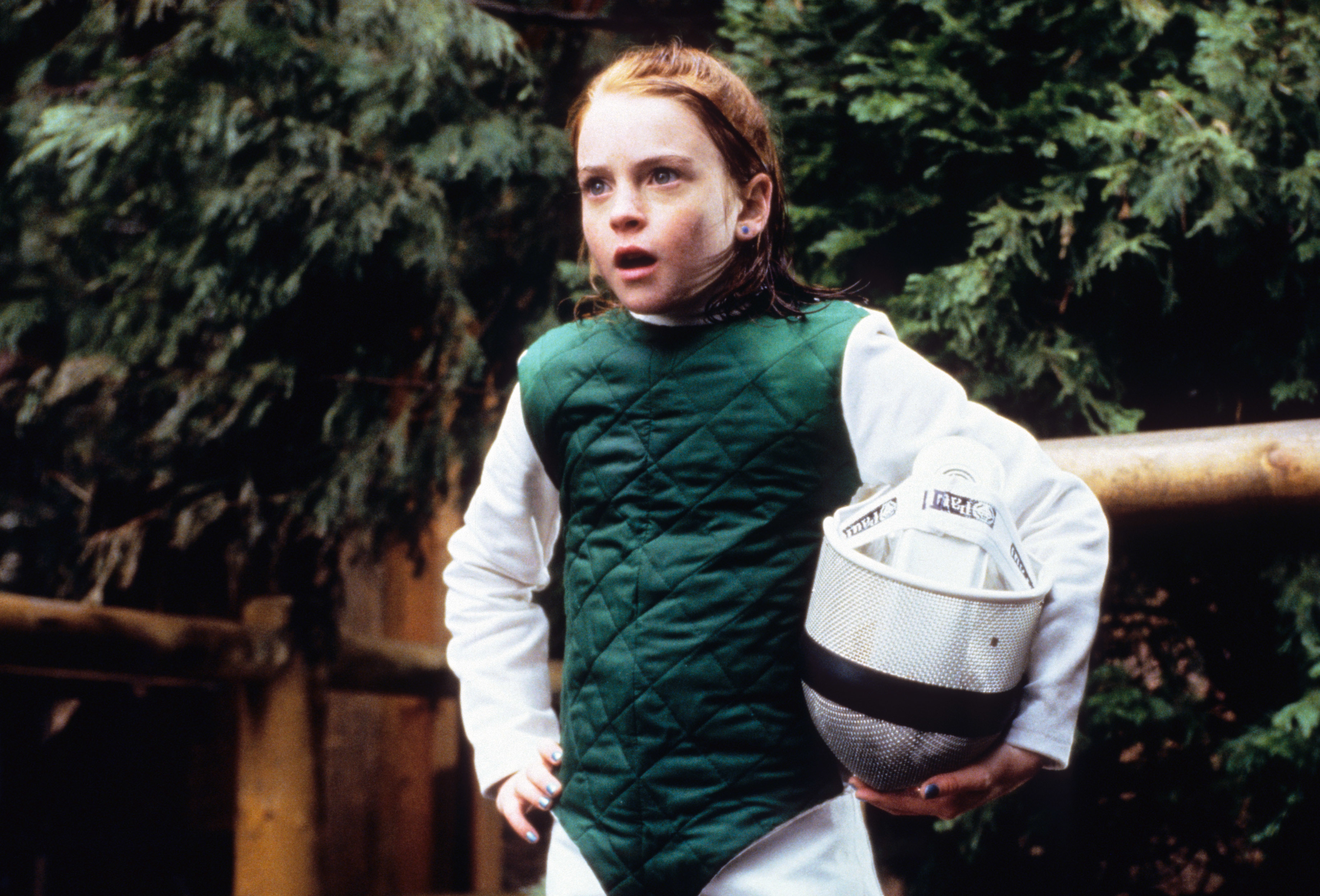 Lindsay Lohan in a fencing outfit, holding a fencing mask under her arm, looks surprised or determined while standing outside in scene from "The Parent Trap"