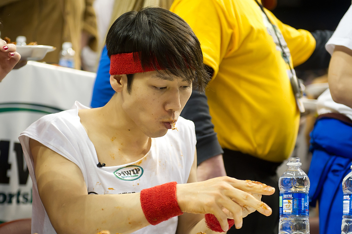 A man wearing a white tank top and red sweatbands is eating a large quantity of chicken wings in what appears to be a competitive eating contest. Other people are in the background