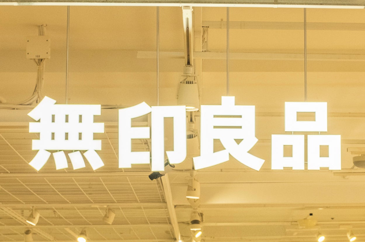 A store interior with shoppers walking around, featuring shelves filled with various items. The store sign has Japanese characters that translate to "MUJI."