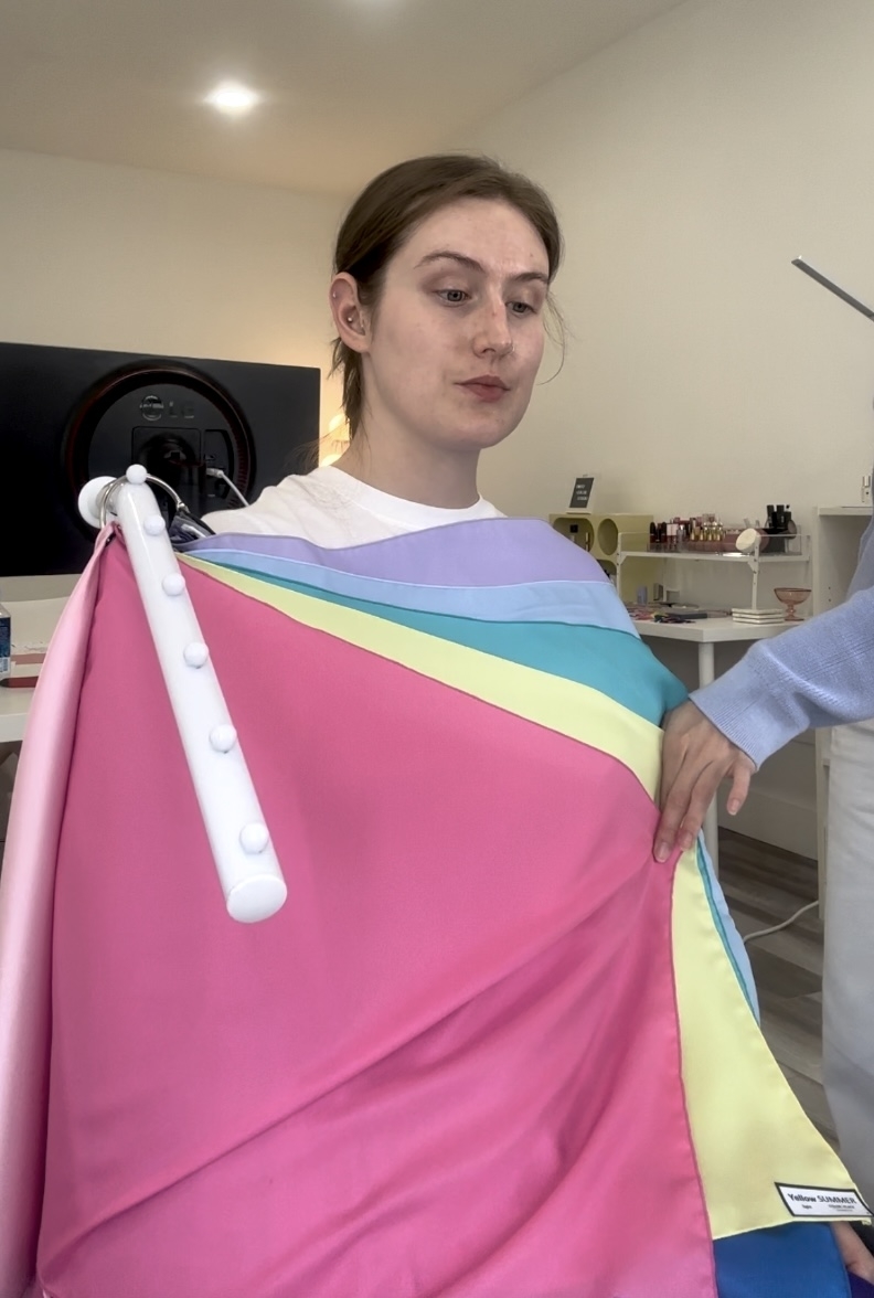 Emily Bridges sits with a multi-colored blanket draped over her, holding it with one hand. She is in a room with a desk and shelves in the background