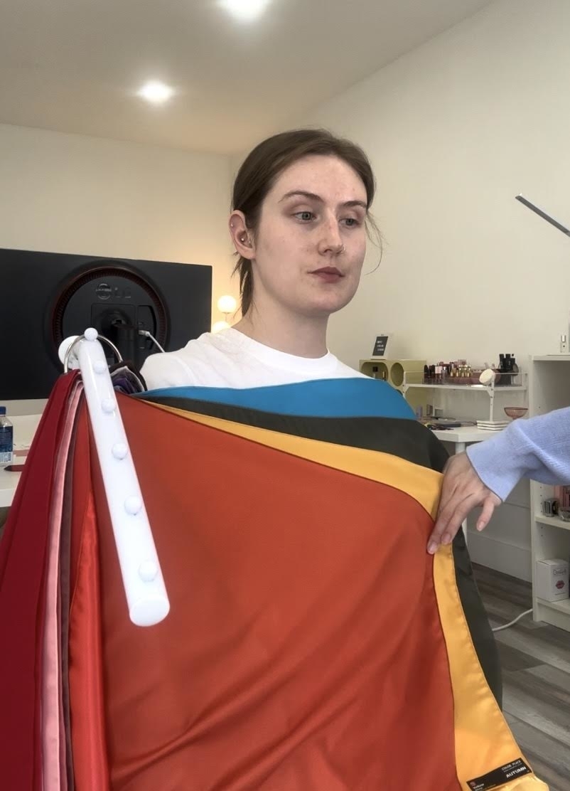 A person stands indoors with various colored fabrics draped around their shoulders while someone holds the fabrics next to them