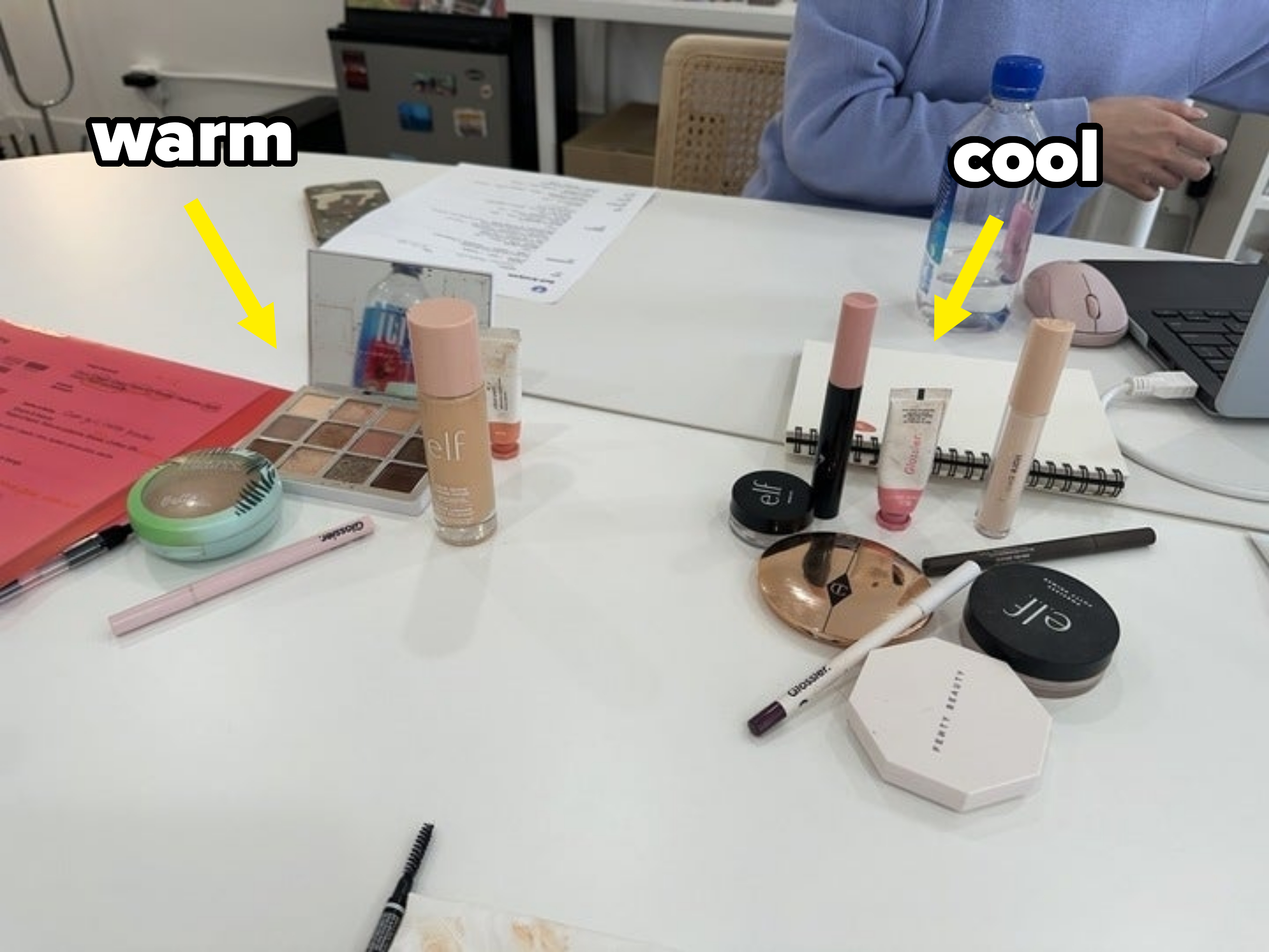 Various makeup products are spread out on a table beside a water bottle, a laptop, and paperwork. A person in a light sweater is partially visible