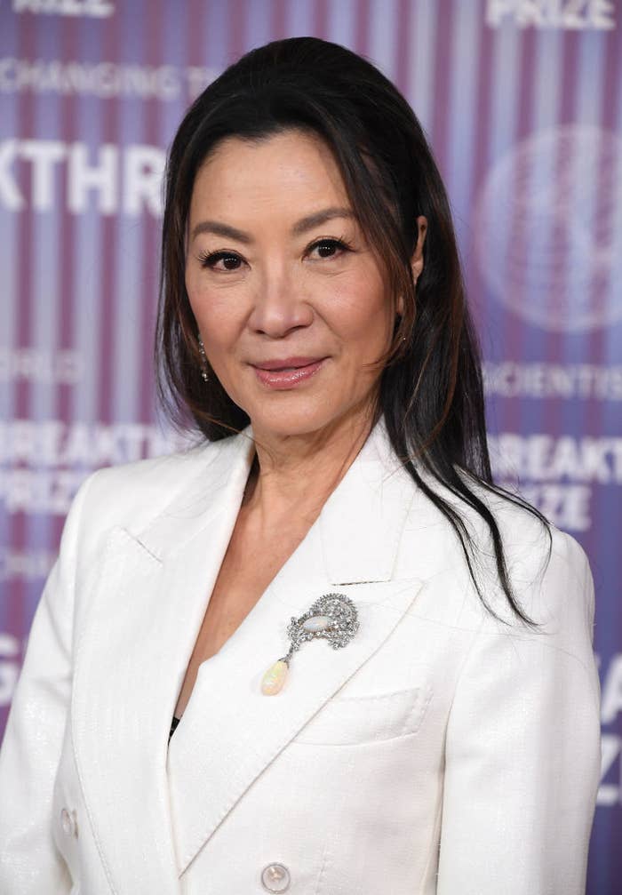 Michelle Yeoh poses elegantly on the red carpet, wearing a white jacket adorned with a detailed brooch