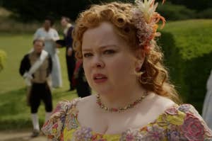 Nicola Coughlan in period costume, featuring curly hair adorned with flowers, a multi-colored floral dress, and a floral necklace, in an outdoor historical setting