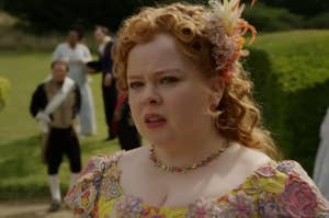 Nicola Coughlan in period costume, featuring curly hair adorned with flowers, a multi-colored floral dress, and a floral necklace, in an outdoor historical setting