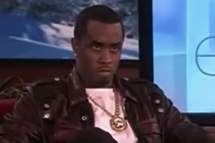 Sean Combs, wearing a jacket with gold buttons and a gold chain necklace, is sitting on a talk show set with a serious expression