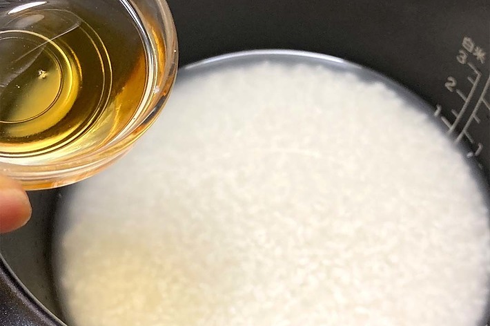 炊飯器にお米と水を入れ、小さなボウルから液体を注ぎ入れる手。