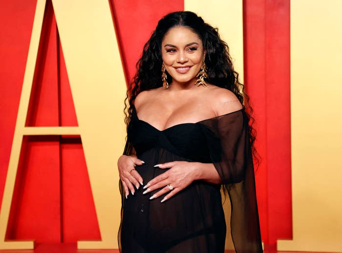 Vanessa Hudgens poses on the red carpet in a sheer dress, holding her baby bump and smiling