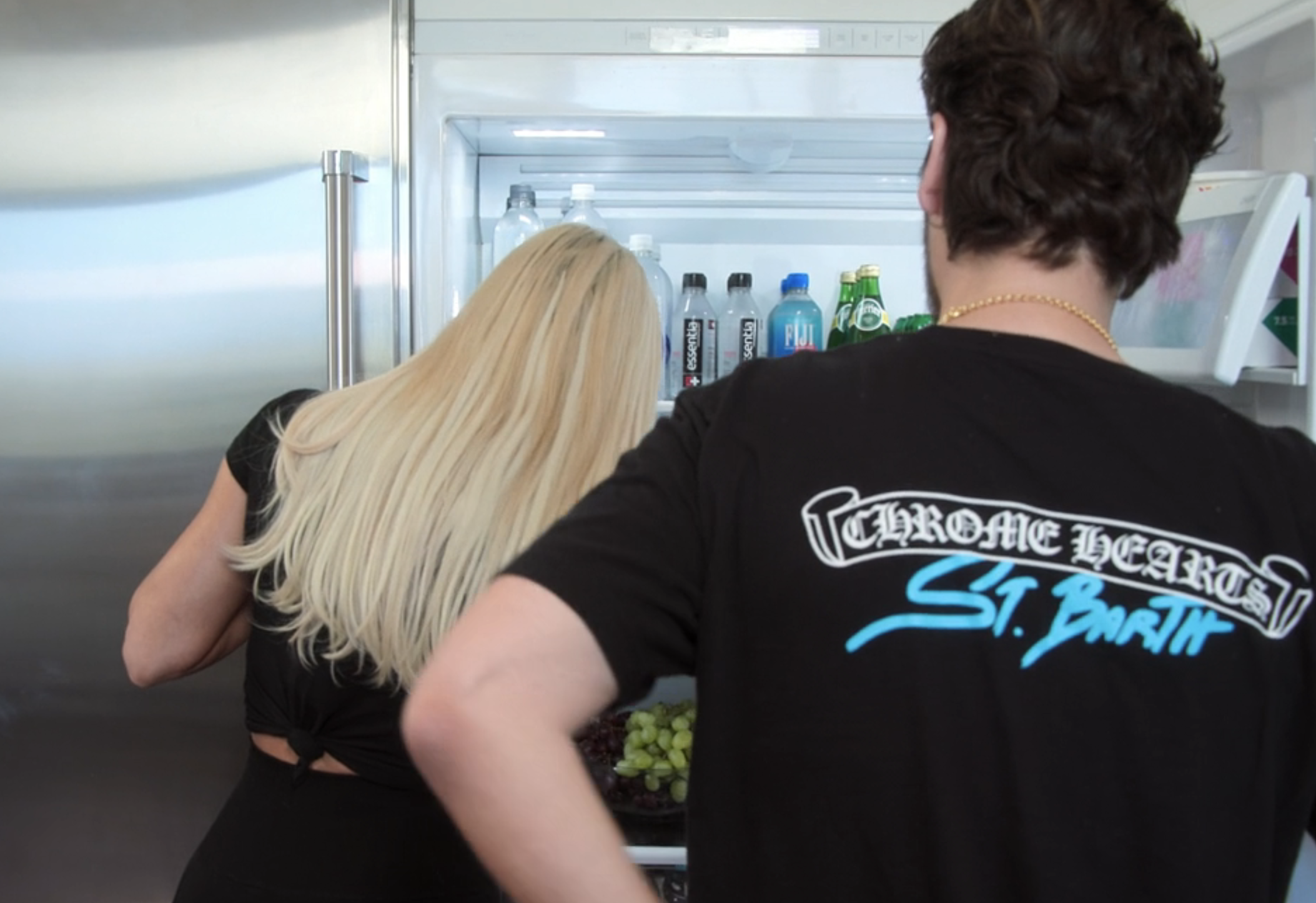 Two people stand in front of an open refrigerator. Person on the right wears a &quot;Chrome Hearts St. Barth&quot; T-shirt. Person on the left has long blonde hair