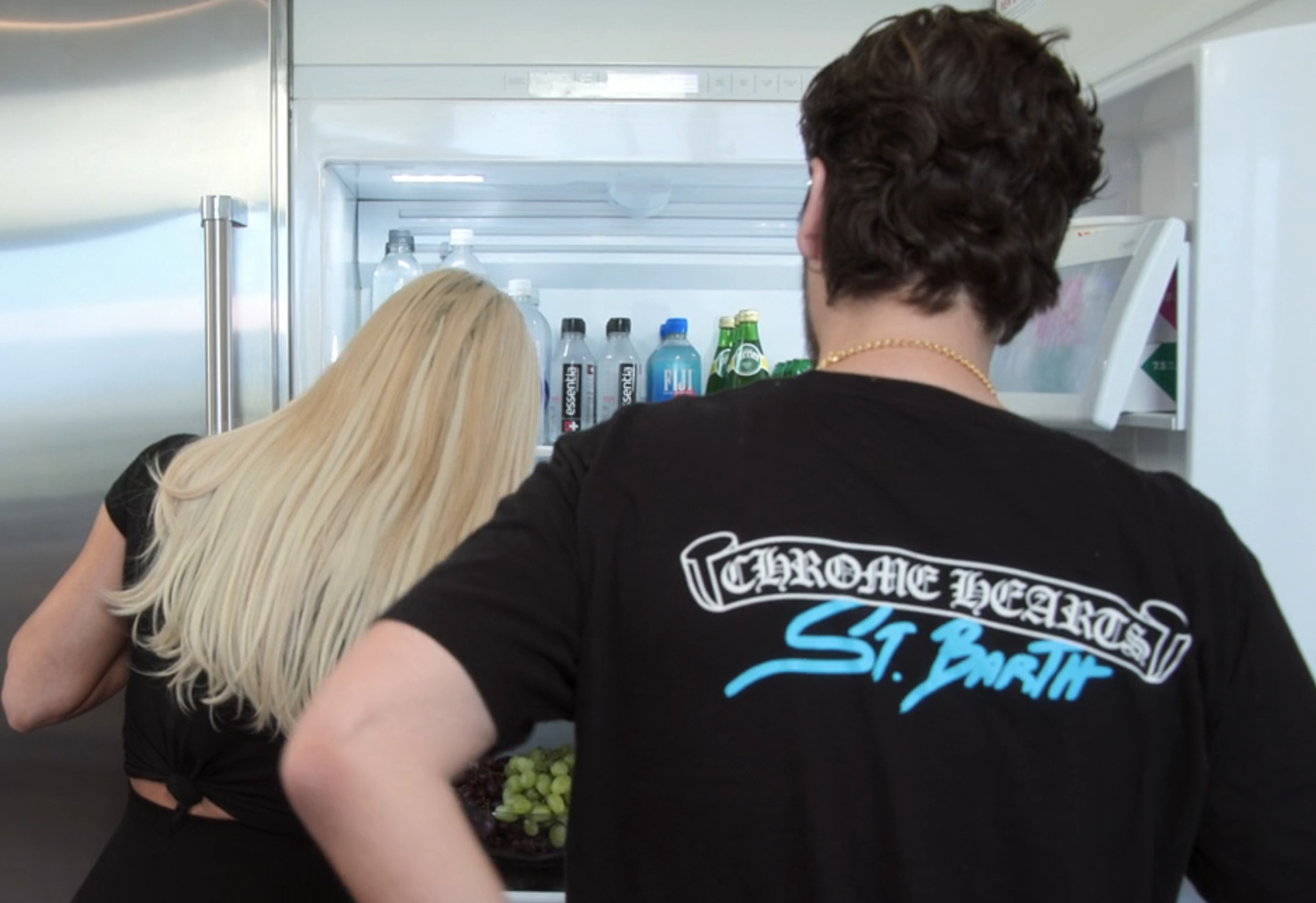 Two people stand in front of an open refrigerator. Person on the right wears a "Chrome Hearts St. Barth" T-shirt. Person on the left has long blonde hair