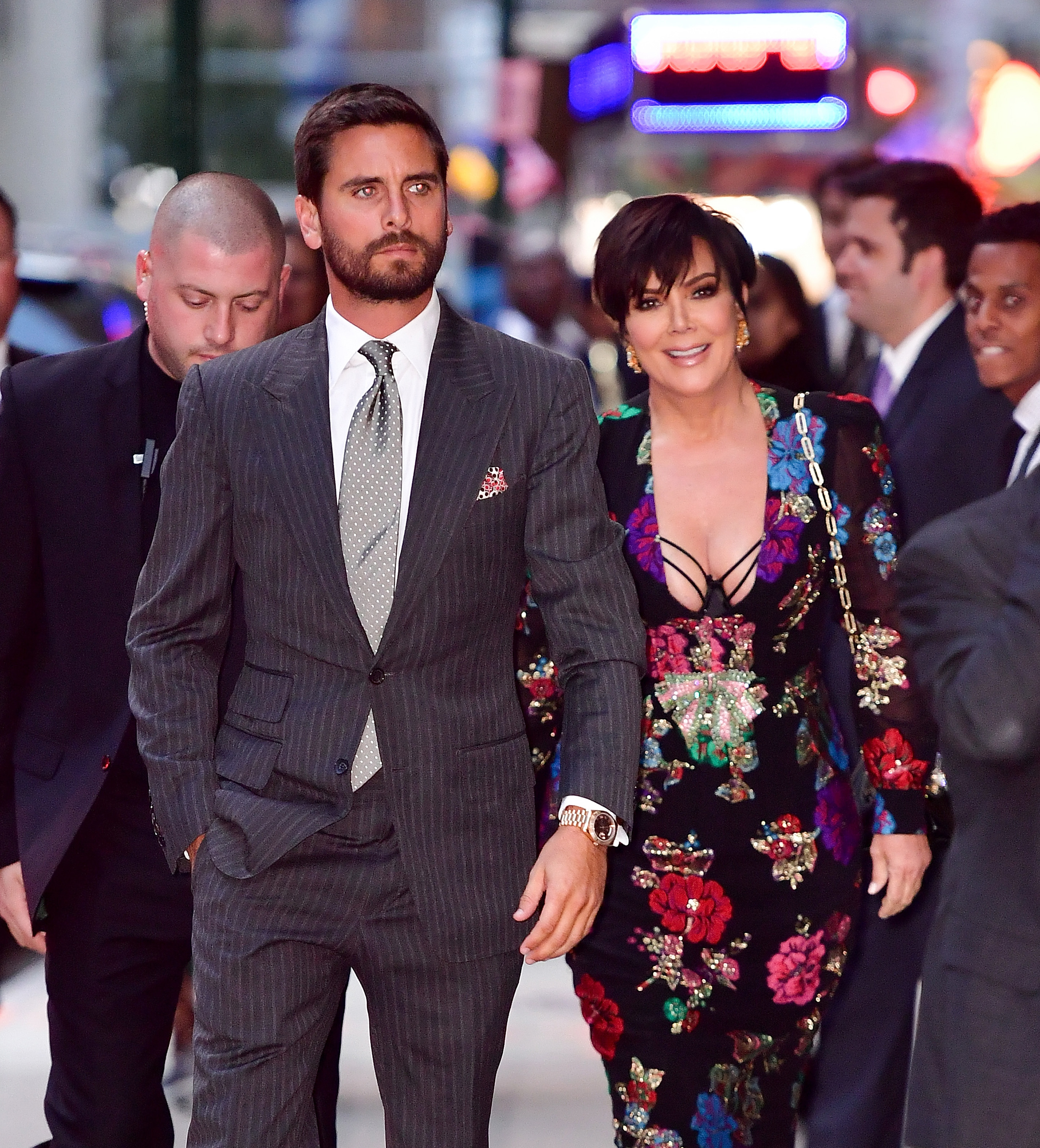 Scott Disick in a pinstriped suit and Kris Jenner in a floral-patterned dress attend a nighttime event, walking among other people in the background