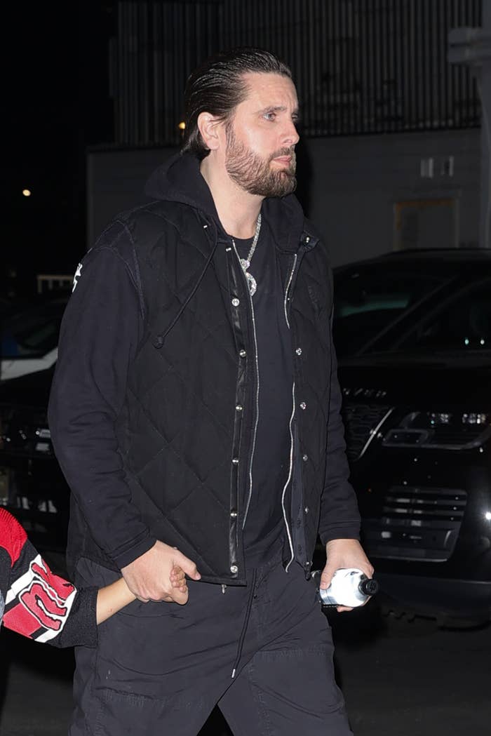 Scott Disick walks, holding a child's hand, wearing a black quilted vest over a hoodie and pants, in front of a dark background with cars