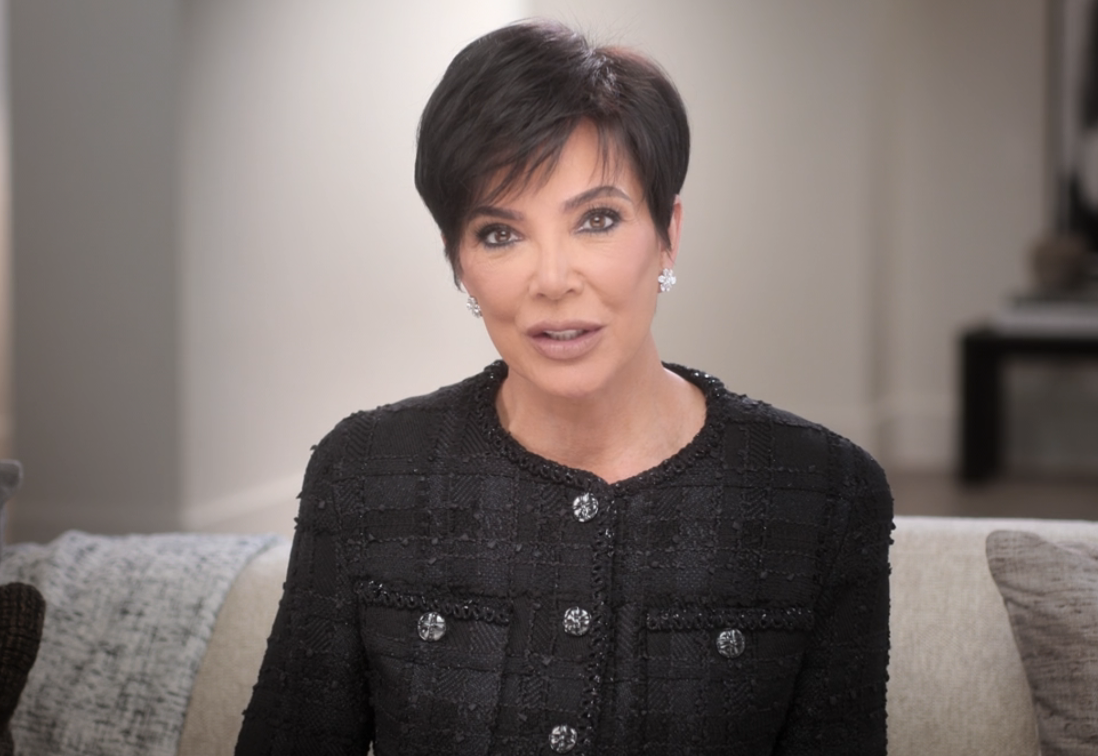 Kris Jenner sits on a couch, wearing a textured, buttoned, black jacket, speaking directly to the camera in a well-lit room. There is an inset of the same image in the bottom-right corner