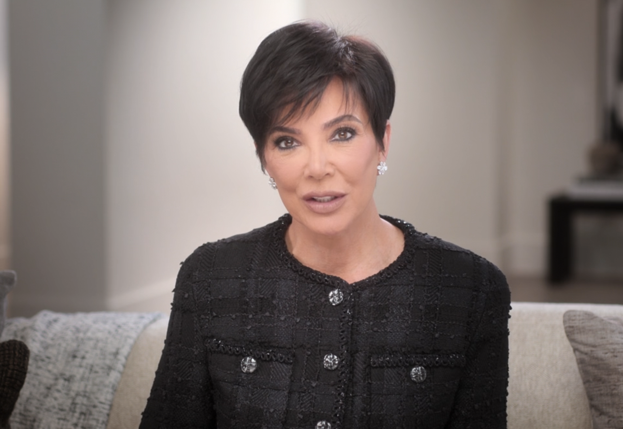 Kris Jenner sits on a couch, wearing a textured, buttoned, black jacket, speaking directly to the camera in a well-lit room. There is an inset of the same image in the bottom-right corner