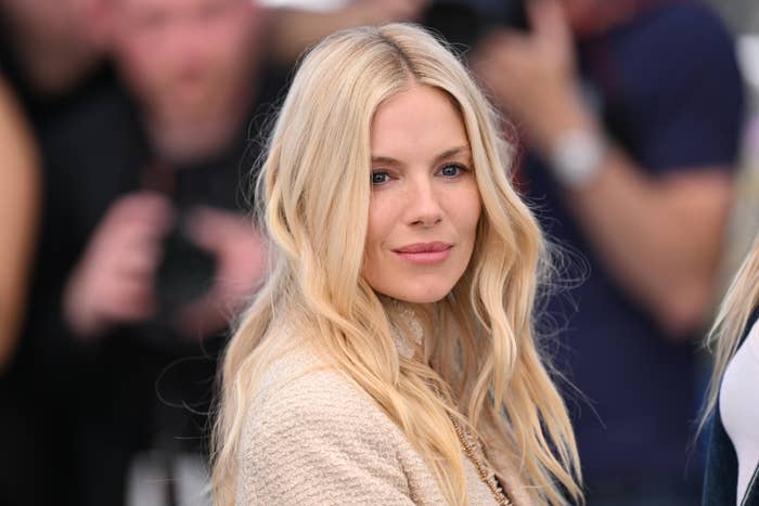 Sienna Miller poses with a neutral expression at a public event. She is wearing a textured, light-colored outfit and her hair is styled in loose waves. Photographers are visible in the background