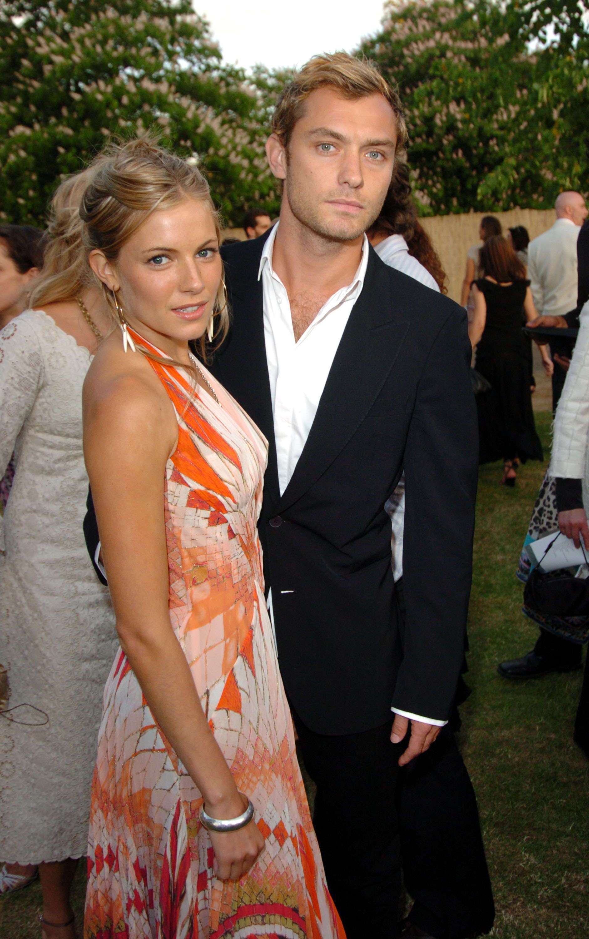 Sienna Miller in a patterned dress and Jude Law in a black suit at an event with outdoor greenery in the background