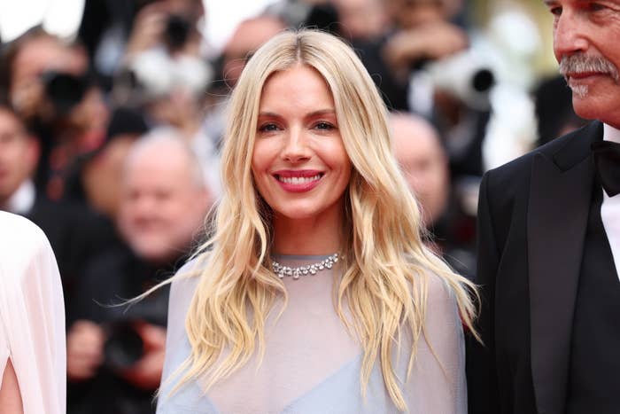 Sienna Miller smiles on the red carpet, wearing a sheer gown with delicate embellishments and a sparkling necklace. She is surrounded by other attendees