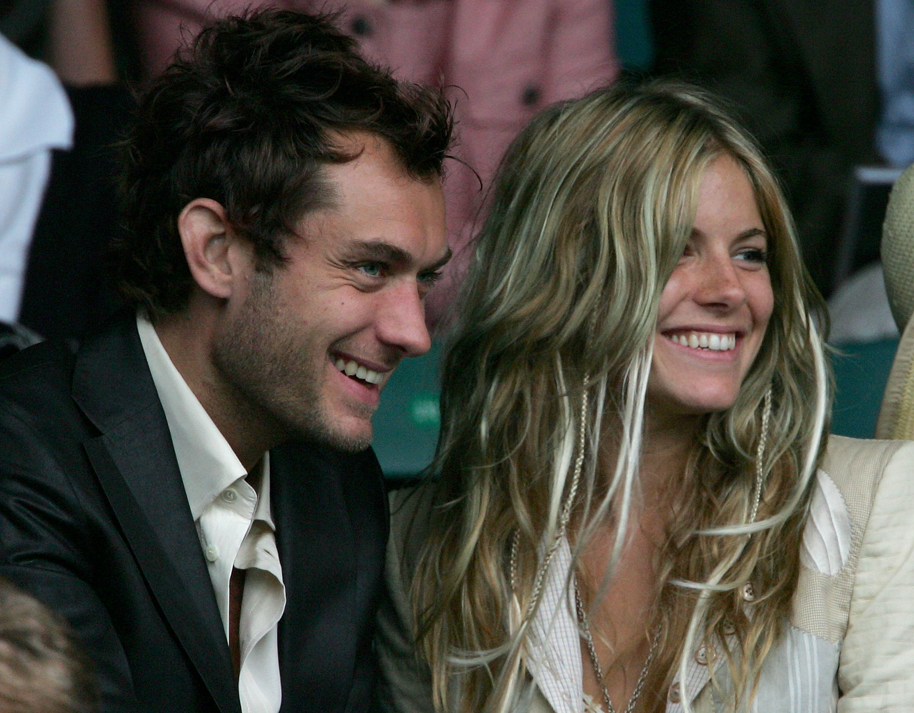 Jude Law and Sienna Miller smiling and sitting close together at an event, both dressed in stylish casual attire