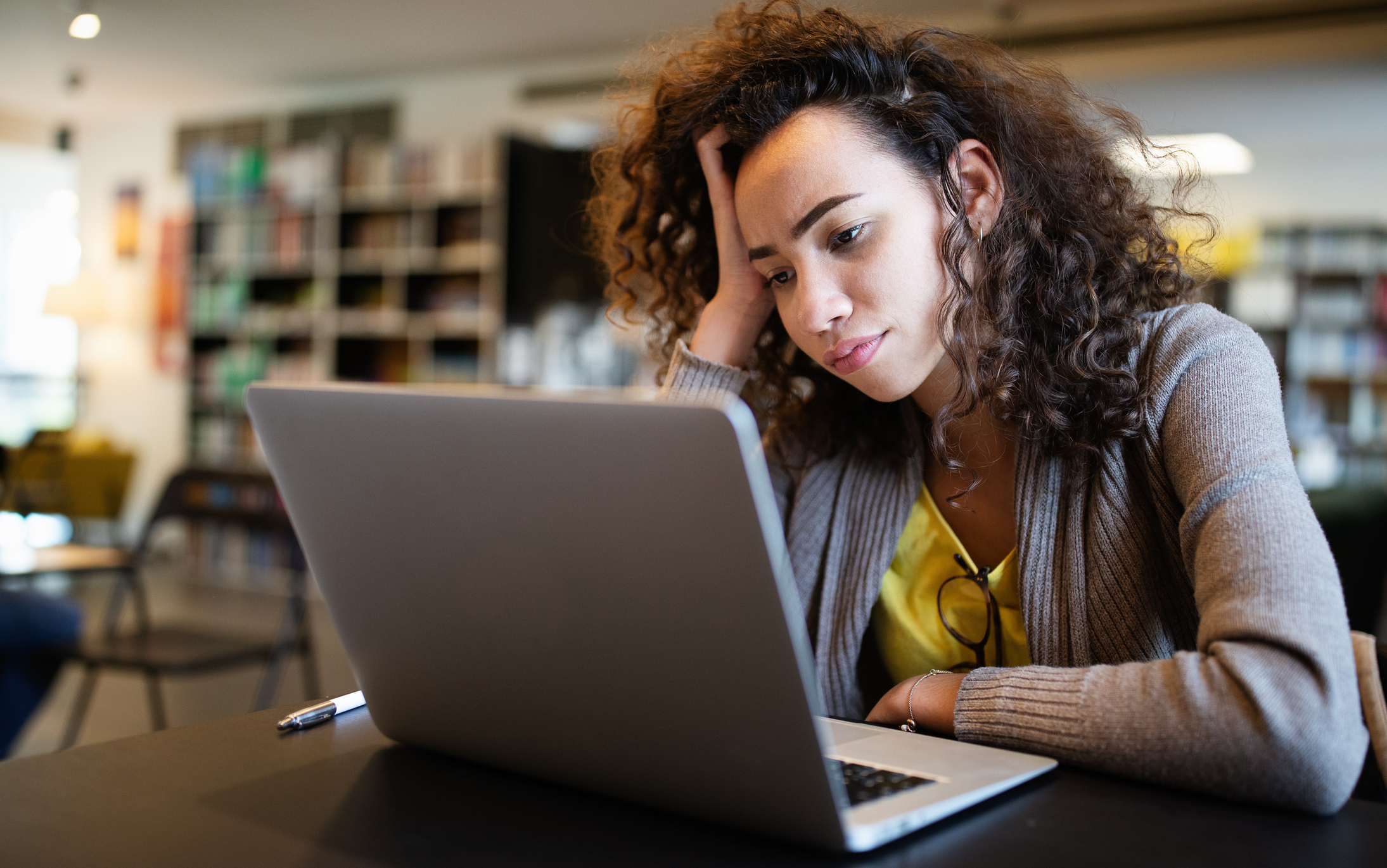 Woman in a sweater sits at a table in a library, looking at her laptop with a thoughtful expression