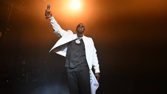 Artist Diddy performing on stage, holding a microphone up, wearing a white jacket, black shirt, and sunglasses, with a spotlight behind him