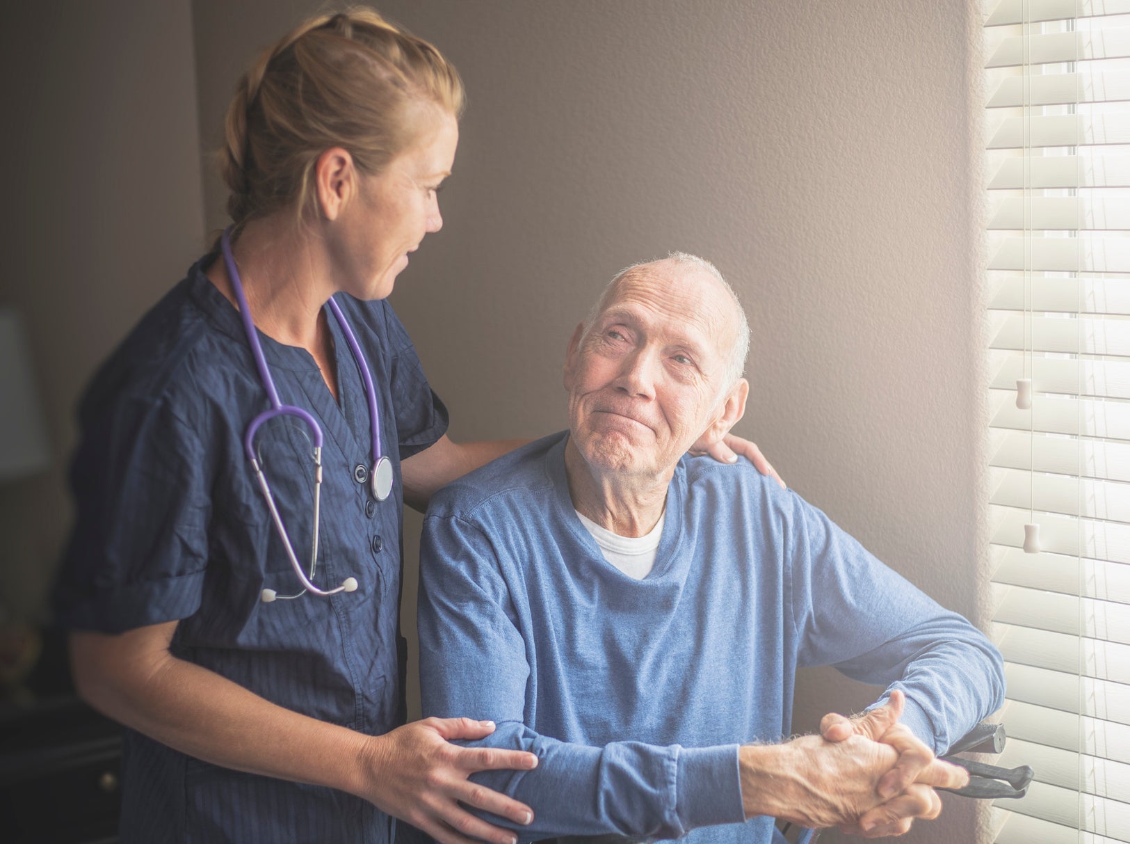 A nurse wearing a stethoscope gently places a hand on the shoulder of an elderly man in a wheelchair, offering comfort and support
