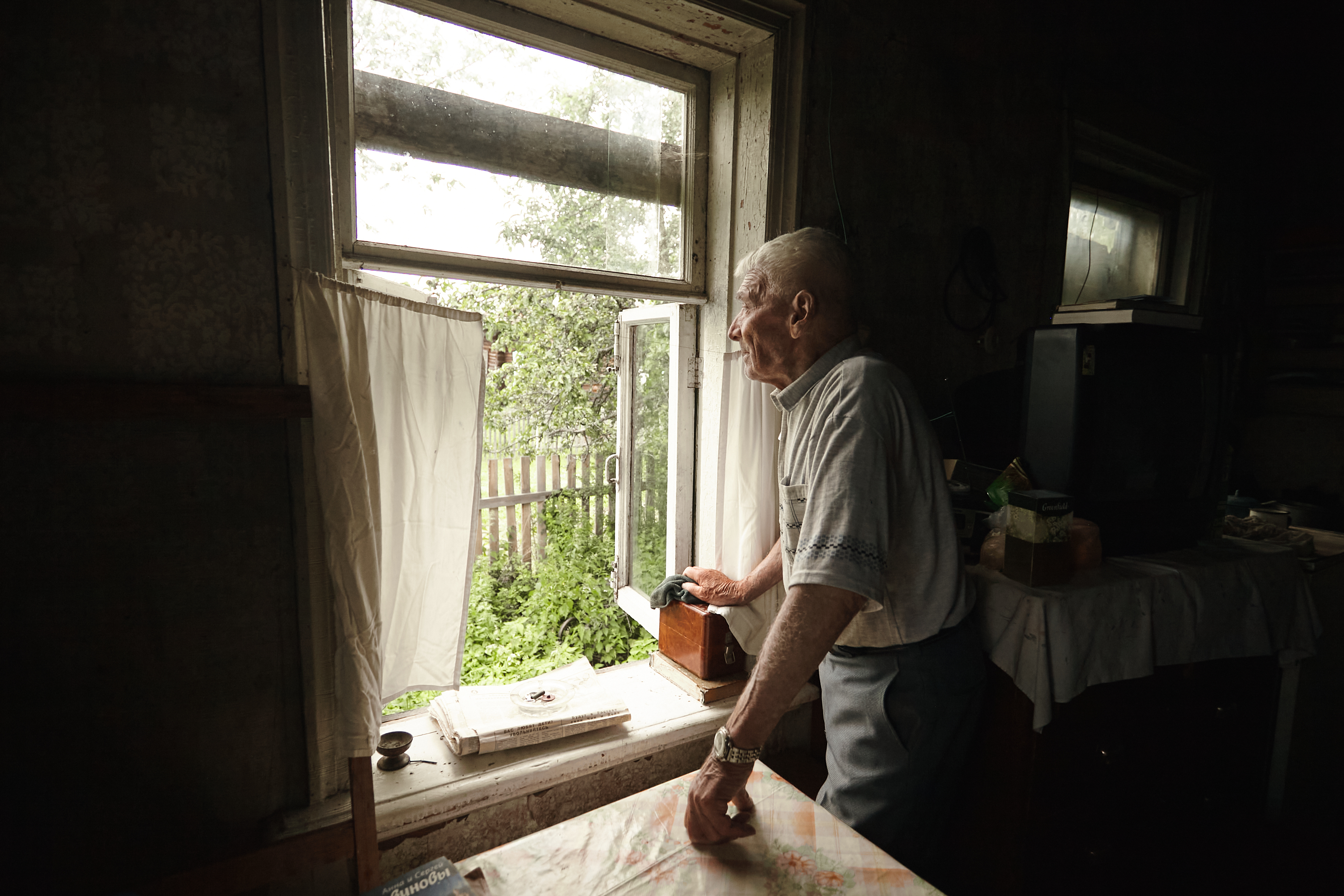 An elderly man stands at a window in a dimly lit room, looking outside thoughtfully while holding a pair of glasses