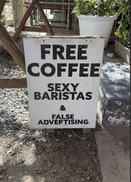 A sign reads, "FREE COFFEE SEXY BARISTAS & FALSE ADVERTISING." The sign is placed on a gravel surface next to a staircase