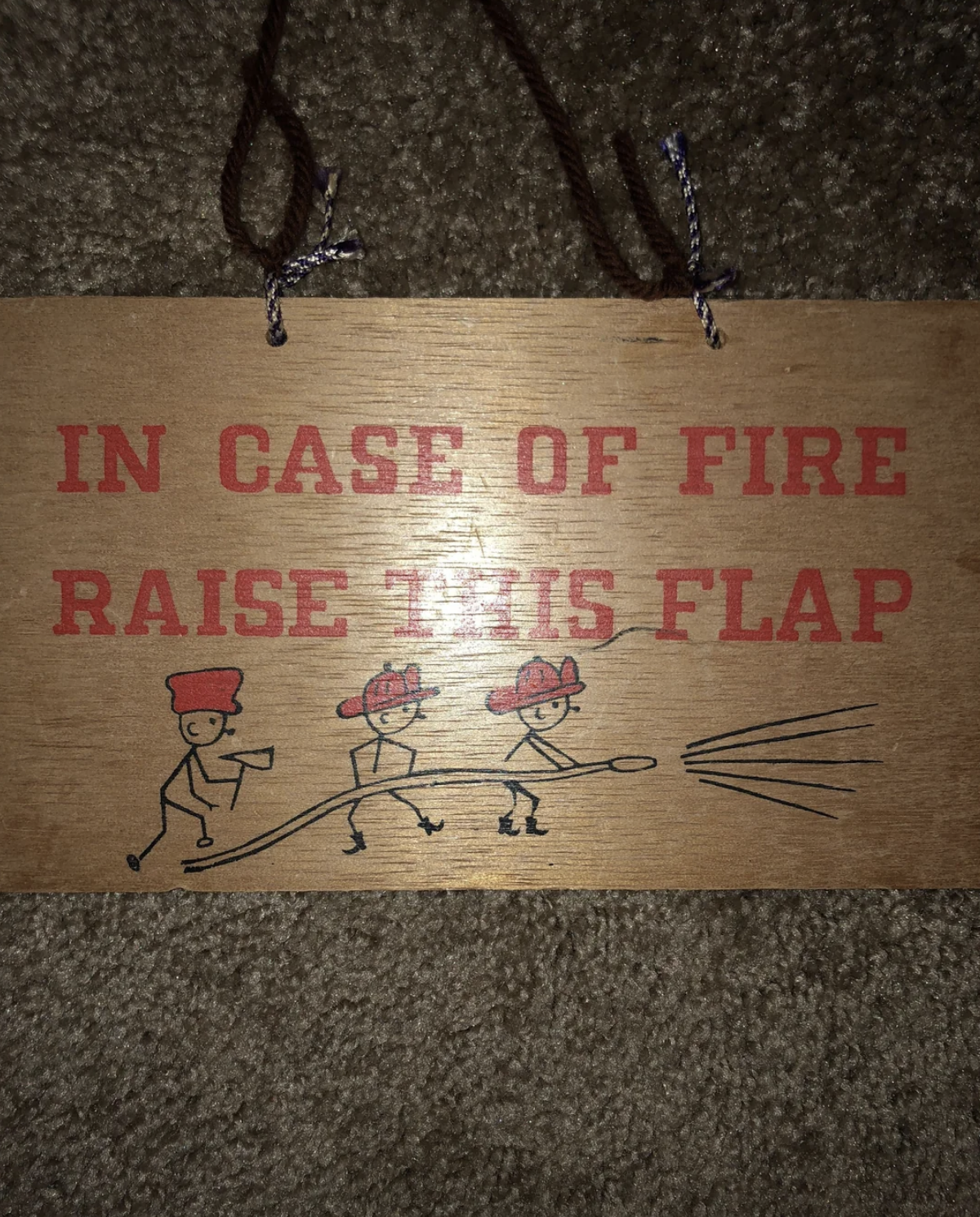 Wooden sign with text "In case of fire raise this flap." Below, three stick figures in fire hats hold a hose spraying water