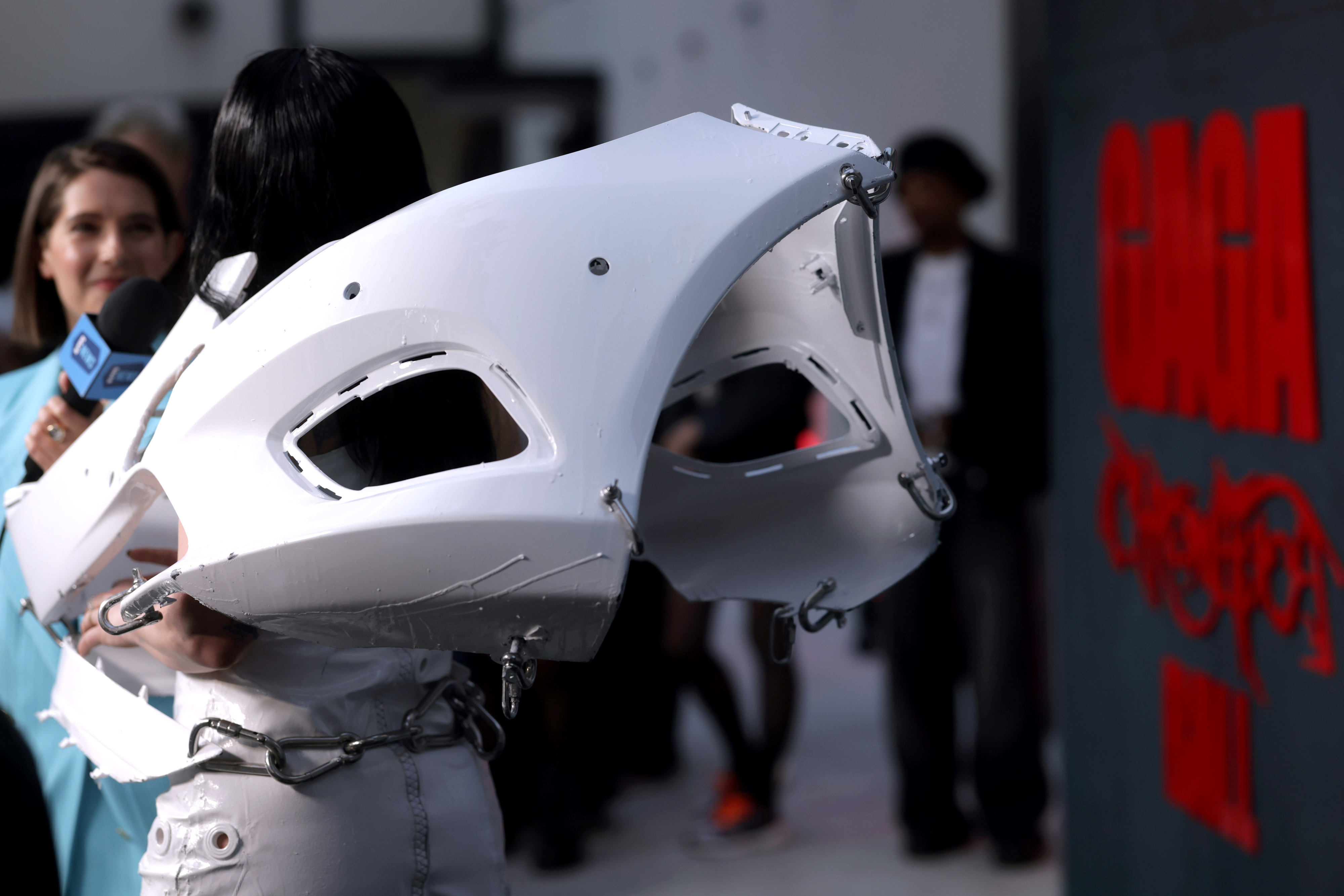 A person is interviewed on the red carpet wearing an avant-garde outfit with large, white sculptural elements. The word &quot;GAGA&quot; is visible in the background