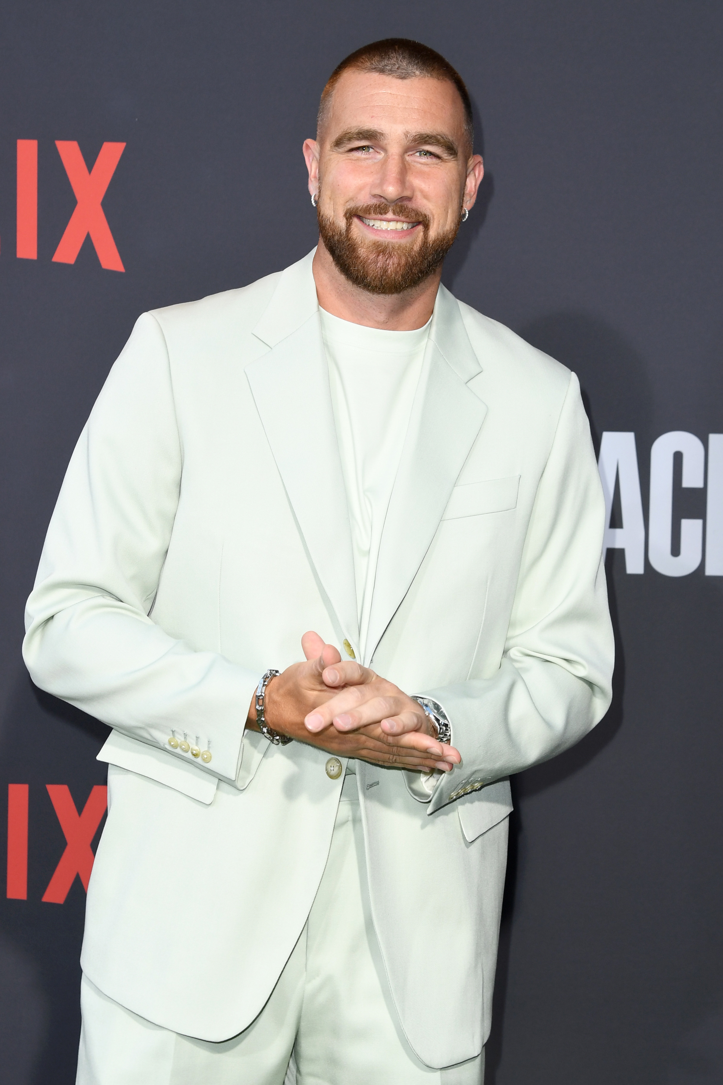 Travis Kelce on the red carpet, wearing a light-colored suit and smiling with hands clasped