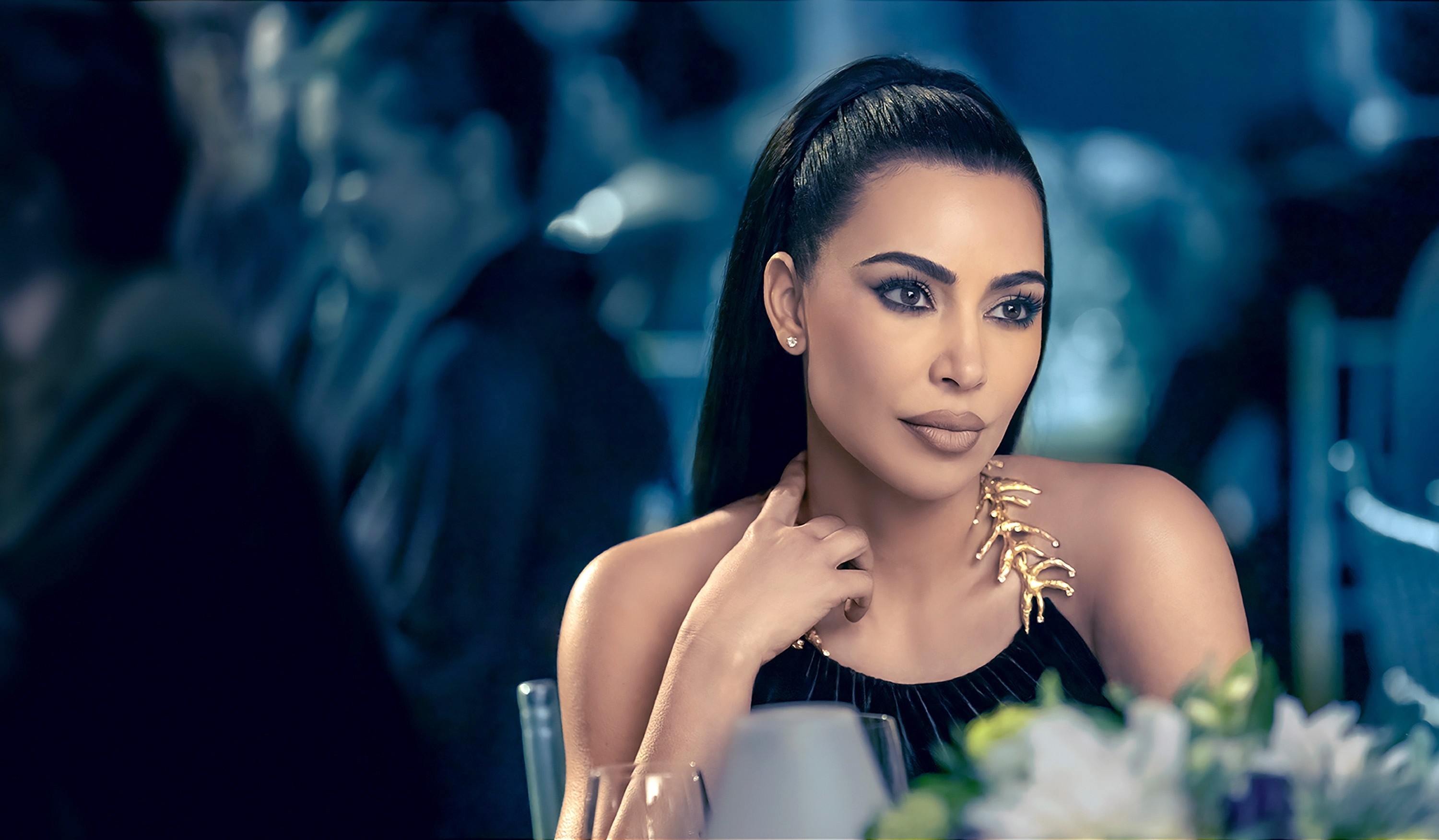Kim Kardashian sits at a table, wearing a sleek outfit with a distinctive gold accessory around her neck. She gazes thoughtfully into the distance in a scene